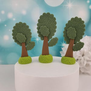 Könnte beinhalten: Drei Miniatur-Baumfiguren. Jeder Baum hat einen braunen Stamm, ein grünes, strukturiertes, rundes Blätterdach und steht auf einer grünen, grasbewachsenen Basis. Die Bäume sind auf einer weißen, runden Oberfläche vor einem verschwommenen, hellblauen Hintergrund angeordnet.