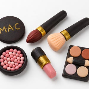 Könnte beinhalten: Ein Set aus Miniatur-Make-up-Artikeln, bestehend aus einem schwarzen Puder-Döschen mit dem goldenen Schriftzug M·A·C, zwei Make-up-Pinseln mit schwarzen Griffen und goldenen Zwingen, einer kleinen Schale mit rosa Kügelchen, einem Lippenstift und einer Palette.