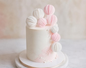 May include: A cylindrical cake with smooth white frosting and a soft pink textured accent at the base. The top and side are decorated with several small, spherical paper honeycomb ornaments in alternating shades of white and pale pink.