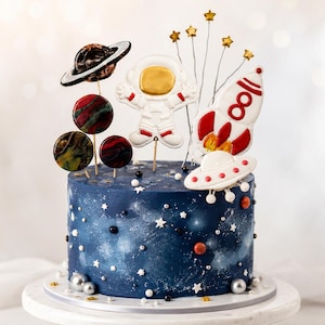 Könnte beinhalten: Ein runder Kuchen mit dunkelblauem Zuckerguss, verziert mit weißen Sternstreuseln und kleinen silbernen Kugeln. Als Dekoration dienen ein Astronautenkeks, eine rot-weiße Rakete, ein UFO, ein Planet mit Ringen und drei runde Planeten.