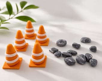 May include: A set of five miniature orange traffic cones with white horizontal stripes and square bases, accompanied by a collection of ten small, smooth, dark gray and black marbled river stones arranged in a cluster on a white surface.