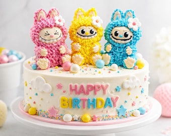 May include: A round white frosted cake topped with three fuzzy-textured monster figures in pink, yellow, and blue. The cake features the text HAPPY BIRTHDAY in colorful letters, decorated with small fondant clouds, stars, and round candy spheres.