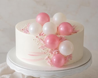 May include: A round cake with smooth white frosting, decorated with a diagonal arrangement of pink and white spherical ornaments, small pink textured flakes, and tiny white pearl sprinkles. The cake sits on a white pedestal stand.