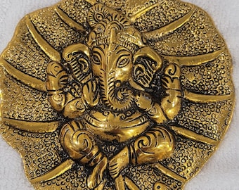 May include: A gold-colored metal wall hanging depicting the Hindu deity Ganesha. The image of Ganesha is centered on a detailed leaf-shaped background. The wall hanging is made of metal and has a textured surface.