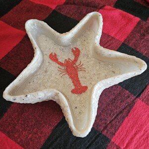 May include: A white starfish-shaped dish with a red glitter lobster design in the center. The dish has a textured surface and is set against a red and black plaid background. This decorative dish is perfect for coastal decor.