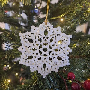 Könnte beinhalten: Ein weißer Schneeflocken-Weihnachtsschmuck mit aufwendigen Ausschnitten, der an einem goldenen Faden hängt. Der Schmuck hängt vor einem grünen Weihnachtsbaum mit Lichtern und roten Beeren.