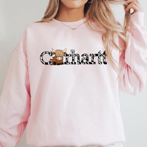 May include: A pale pink sweatshirt with the word "Carhartt" in a black font with a cow print pattern. A cartoon Highland cow is incorporated into the design. The sweatshirt is a plain, light pink colour.