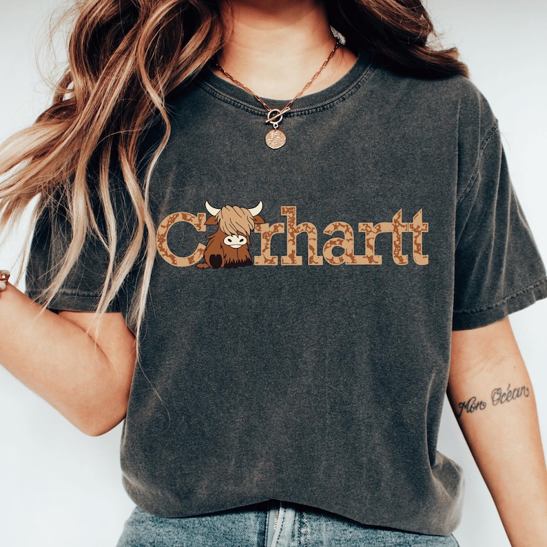 May include: Dark grey t-shirt with the word "Carhartt" in a brown, patterned font, and a cartoon Highland cow graphic. The shirt has a crew neck and short sleeves. The style is casual and comfortable.