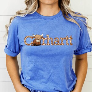 May include: A blue t-shirt with the word "Carhartt" in a brown and white cow print font. A cartoon Highland cow illustration is part of the design. The sleeves are rolled up.