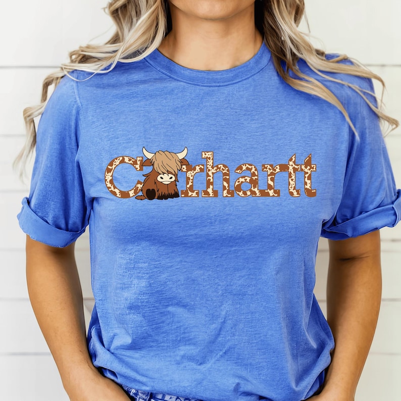 May include: A blue t-shirt with the word "Carhartt" in a brown and white cow print font. A cartoon Highland cow illustration is part of the design. The sleeves are rolled up.