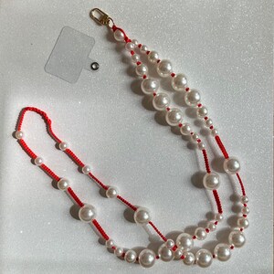 May include: A red string necklace with white pearl beads. The necklace has a gold clasp and a small tag attached to it.