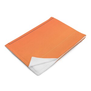 Puede incluir: Una manta suave de color naranja con un reverso blanco. La manta está doblada por la mitad y el lado naranja está hacia arriba.