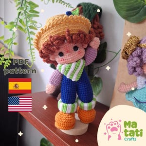 Puede incluir: Una muñeca de ganchillo con un sombrero amarillo estilo paja, pelo castaño rizado, una bufanda a rayas verdes y blancas, una camiseta morada de manga larga, un peto azul y zapatos naranjas. La imagen incluye el texto PDF pattern, una bandera española y una estadounidense.