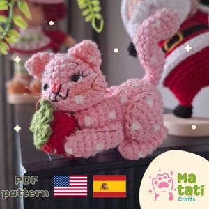 May include: A pink crocheted cat with white polka dots, holding a red strawberry with green leaves. The cat has a smiling face and whiskers. The image also includes the text "PDF pattern" and the American and Spanish flags.