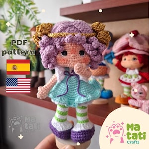 May include: A crochet doll with curly purple hair, gold accents, and a light blue dress. The doll wears white and green striped socks and purple shoes. Text on the image includes PDF pattern, the Spanish flag, the American flag, and Ma tati Crafts.
