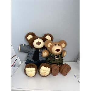 Op de afbeelding: Twee pluche teddyberen. Eén beer is gekleed in een grijs uniform met een zwarte stropdas, de andere in een camouflage-outfit. Beide beren hebben bruine vacht, crèmekleurige snuiten en grote, ronde voeten. De beer links heeft de naam "Jedgar" op zijn voet.