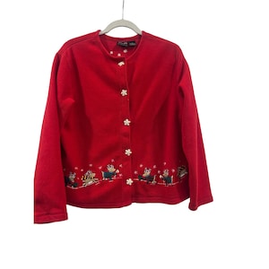 Peut inclure: Un cardigan rouge avec un col rond et des manches longues. Le pull est orné de boutons en forme de flocons de neige et d'une broderie de rennes et de flocons de neige en bas. Le pull est suspendu à un cintre blanc.
