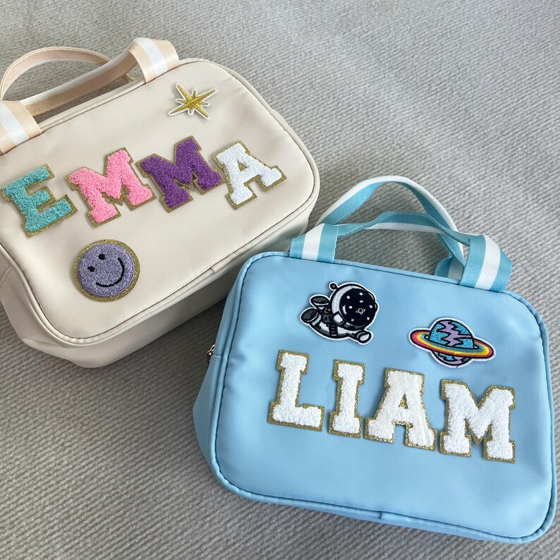 May include: Two personalized lunch bags, one cream colored with the name "EMMA" in colorful letters and a smiley face patch, and one light blue with the name "LIAM" in white letters and astronaut and planet patches.