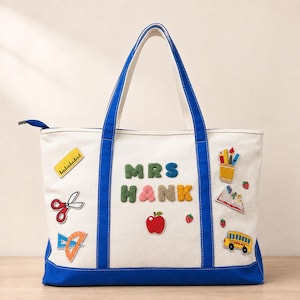 May include: A cream-colored canvas tote bag with blue trim and handles, featuring the text MRS HANK in colorful fuzzy letters. The bag is decorated with embroidered patches of a ruler, scissors, protractor, apple, school bus, and school supplies.