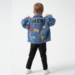 May include: A blue denim jacket with the name "JACK" in black lettering on the back. The jacket is adorned with patches of a football, a dinosaur, and various vehicles. The person is wearing black jeans. The jacket is made of denim.