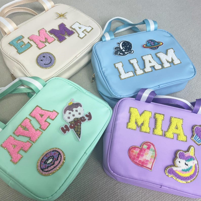 May include: Four personalized lunch bags with different colors and designs. The bags are made of a soft, durable material and feature a zipper closure. Each bag has a different name on it, including Emma, Liam, Ava, and Mia. The bags are decorated with various patches, including a smiley face, a planet, an ice cream cone, a donut, a heart, and a unicorn.