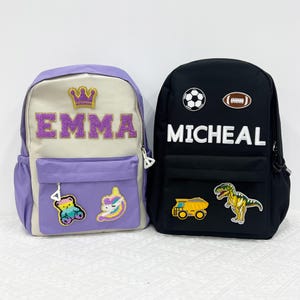 Peut inclure: Deux sacs à dos. L'un est lavande et crème avec le nom "EMMA" en lettres pailletées, une couronne dorée et des écussons d'ours et de licorne. L'autre est noir avec "MICHEAL" en lettres blanches, des écussons de ballon de foot, de football américain, de camion benne et de dinosaure.
