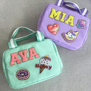May include: Two personalized lunch bags, one green with the name "AVA" and patches of a donut, ice cream cone, and a heart, and one purple with the name "MIA" and patches of a cupcake, unicorn, and a heart.
