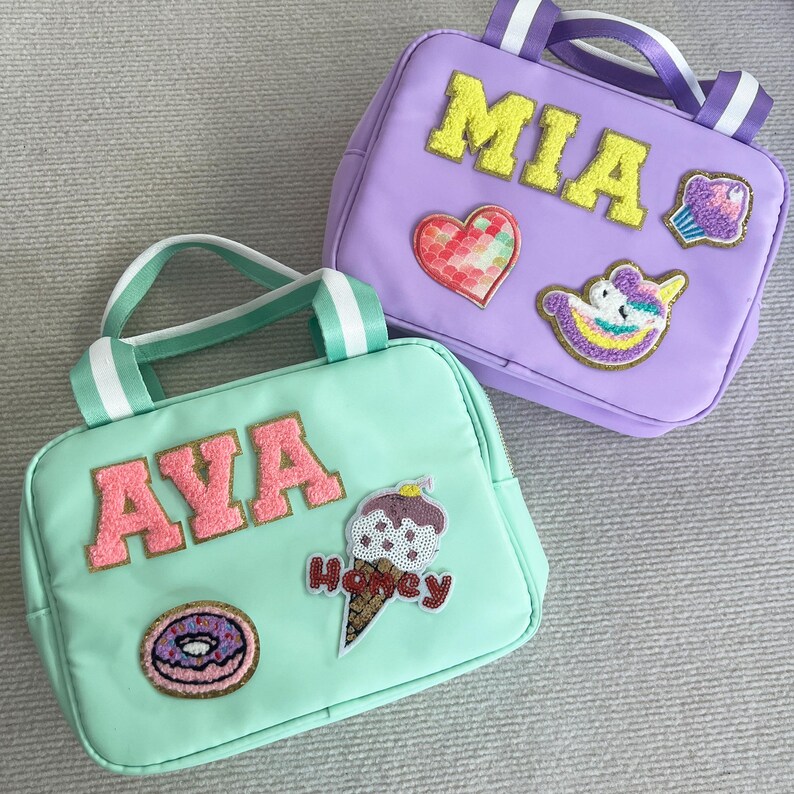 May include: Two personalized lunch bags, one green with the name "AVA" and patches of a donut, ice cream cone, and a heart, and one purple with the name "MIA" and patches of a cupcake, unicorn, and a heart.