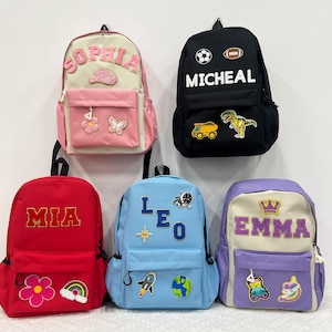 Peut inclure: Cinq sacs à dos colorés avec des noms personnalisés et des écussons décoratifs. Les sacs à dos sont de différentes couleurs, dont le rouge, le noir, le bleu clair, le rose et le violet. Chaque sac à dos porte un nom différent, comme "MIA", "MICHEAL", "SOPHIA", "LEO" et "EMMA".