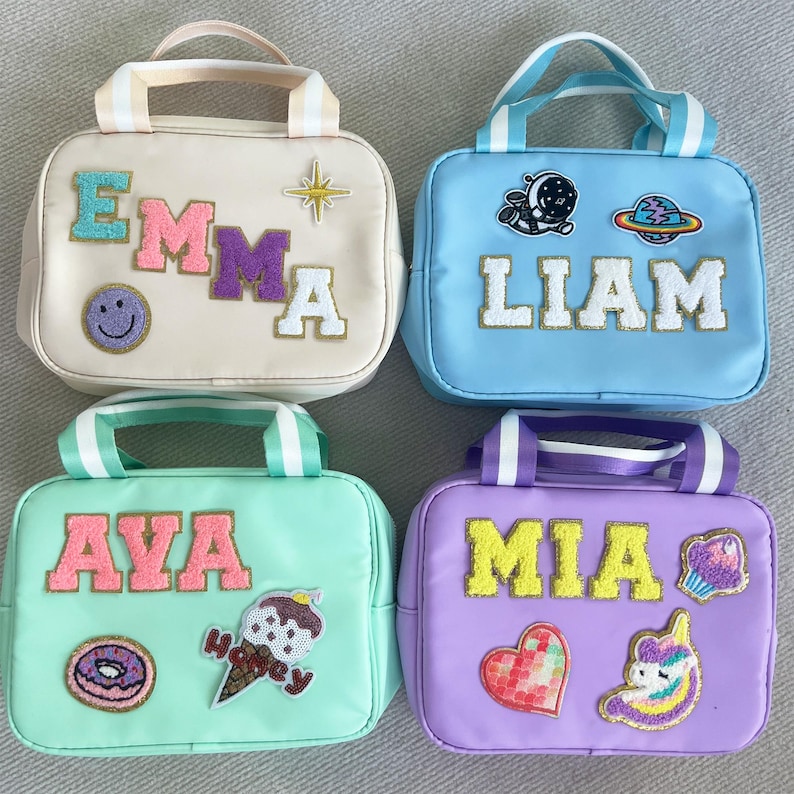 May include: Four personalized lunch bags with different names and colorful patches. The bags are beige, blue, green, and purple. The names on the bags are Emma, Liam, Ava, and Mia.