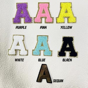 May include: Six iron-on patches in different colors and textures, each featuring a large capital letter "A". The colors are purple, pink, yellow, white, blue, and black. The last patch is gold and has a sequin texture. The patches are labeled with their colors below.