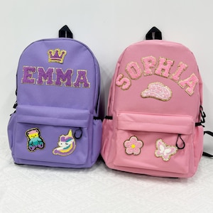 Peut inclure: Deux sacs à dos, un lavande et un rose, sont présentés. Le sac à dos lavande porte le nom "EMMA" en lettres pailletées et une couronne. Le sac à dos rose porte le nom "SOPHIA" en lettres pailletées et un nuage. Les deux ont des poches avant.