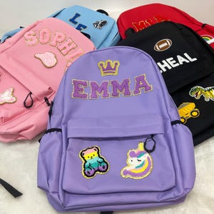 Peut inclure: Plusieurs sacs à dos colorés avec des écussons brodés et du texte. Le sac à dos violet central porte le nom "EMMA" en lettres pailletées, une couronne dorée et des écussons d'ours en peluche et de licorne. D'autres sacs sont roses, bleus, rouges et noirs.