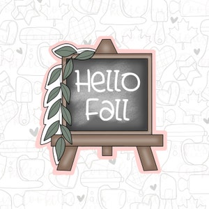 May include: A brown wooden easel with a chalkboard that says "Hello Fall" in white text. The easel is decorated with a green leafy garland.