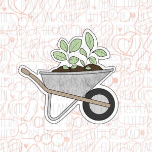 May include: A cookie cutter in the shape of a wheelbarrow filled with soil and small plants. The wheelbarrow is silver with a brown handle and a black wheel. The plants have green leaves.