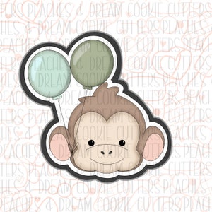 May include: A cookie cutter in the shape of a monkey holding two balloons. The monkey is brown with a light brown face and pink ears. The balloons are green and light blue.