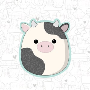 May include: A cute cartoon cow with black and white spots and a pink nose. The cow is surrounded by a light blue outline and is set against a background of white with gray outlines of kitchen tools.