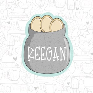May include: A grey cookie cutter shaped like a jar with a lid, filled with gold coins. The jar has the name "KEEGAN" written on the front.