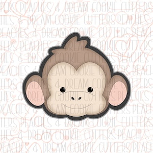 May include: A brown monkey face cookie cutter with a light brown face, pink ears, and a small black nose. The cookie cutter is on a white background with a pattern of pink hearts and lines.