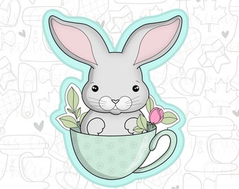 May include: A gray bunny rabbit sits in a light blue teacup with a green floral pattern. The bunny has pink ears and a pink rose behind it.