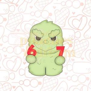 May include: A light green cookie cutter shaped like a cartoon monster holding the numbers 6 and 7 in red. The monster has a smiling face with brown eyes and a jagged outline. The background is a light pink pattern.