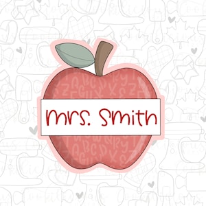 May include: A red apple with a green leaf and a white banner that says "mrs. smith" on a white background with a pattern of various kitchen tools and shapes.