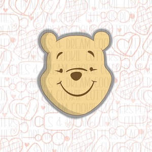 May include: A Winnie the Pooh cookie cutter with a smiling face. The cutter is made of metal and has a gray outline. The background is a white pattern of hearts and other cookie cutters.