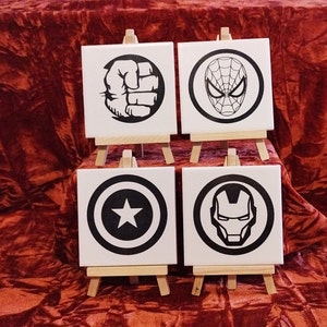 May include: Four square white tiles, each featuring a different black superhero logo: a fist, Spider-Man's mask, Captain America's shield, and Iron Man's helmet. The tiles are on small wooden easels, against a red velvet backdrop.