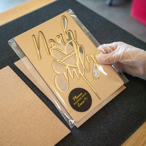 May include: A brown cardboard cake topper with a gold glitter design that says "Noah & Emily" with a heart and the date "10.22.2023". The topper is packaged in a clear plastic bag with a black circle sticker that says "Please handle with love".