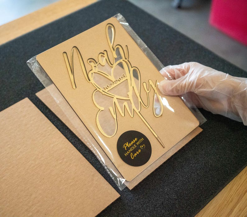 May include: A brown cardboard cake topper with a gold glitter design that says "Noah & Emily" with a heart and the date "10.22.2023". The topper is packaged in a clear plastic bag with a black circle sticker that says "Please handle with love".