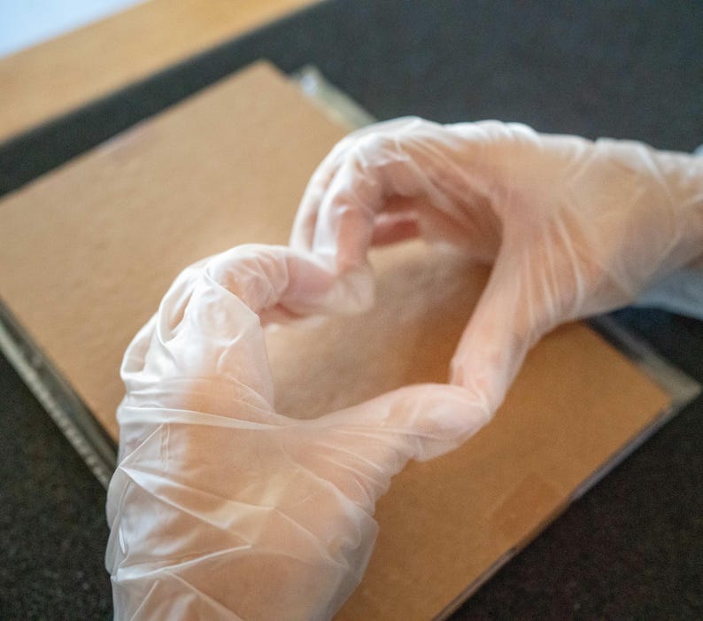 May include: Two hands wearing white latex gloves are making a heart shape on a brown surface.