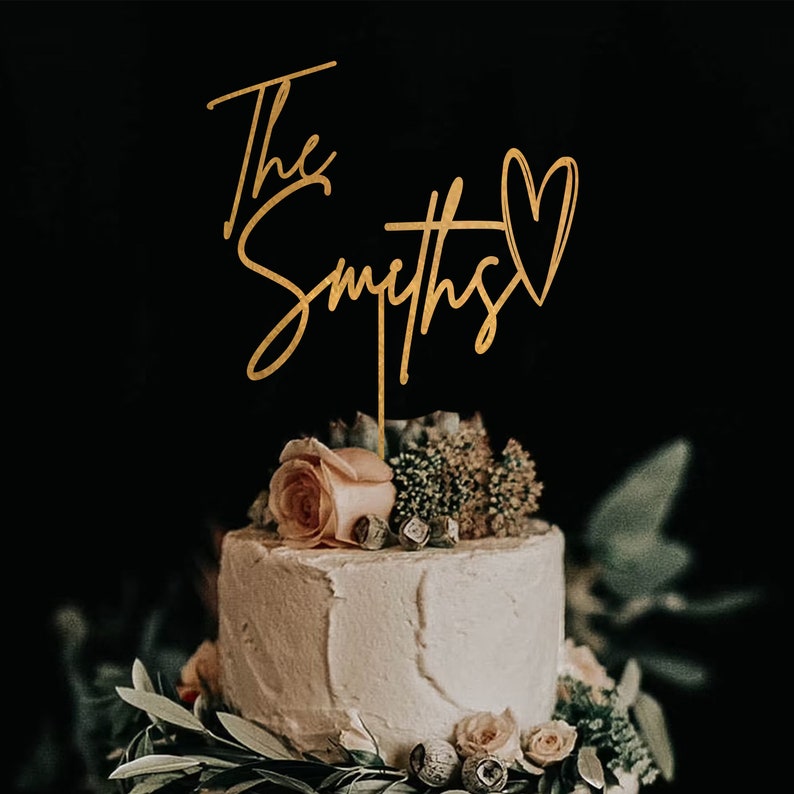 May include: Gold cake topper with the text "The Smiths" and a heart. The topper is on a white cake with floral decorations.
