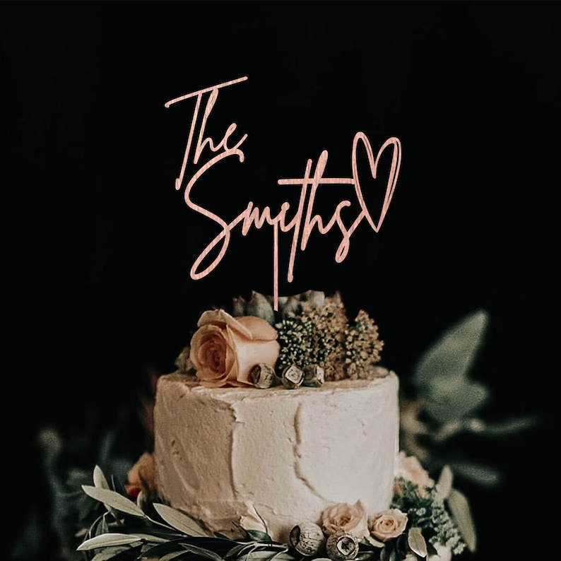 May include: A two-tiered wedding cake decorated with greenery and a single rose. The cake is topped with a pink acrylic cake topper that reads "The Smiths" with a heart.