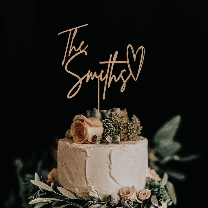 May include: A wedding cake topper featuring the words "The Smiths" in cursive script and a heart symbol. The topper is crafted from wood and is a light brown colour.